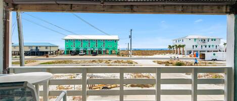 A beachside view from a covered porch with a white railing, overlooking a road with buildings, including a green house and a white house with palm trees.