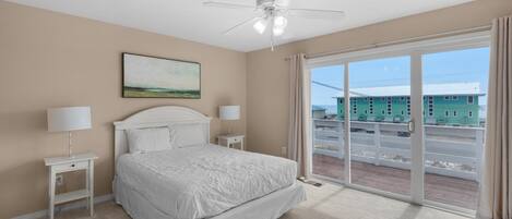A bedroom with a bed, nightstands, a ceiling fan, a painting, and a view of a building through a sliding glass door.