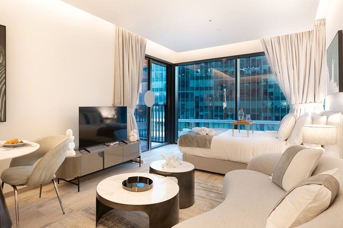 A modern hotel room with a large window, a sofa, a TV, a dining area, and a small table.