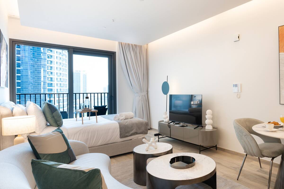 A modern hotel room with a bed, a TV, a dining area with a table and chairs, and a large window with a city view.
