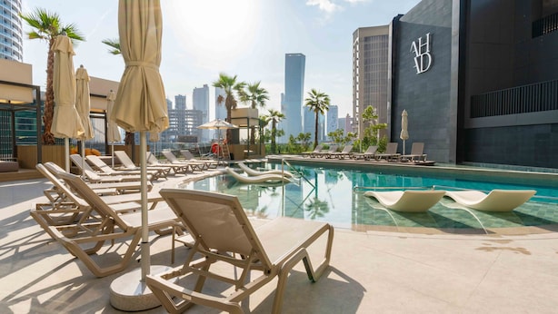 A rooftop pool area with lounge chairs and umbrellas, surrounded by palm trees and modern buildings. - Ahad Residences (Dubai)