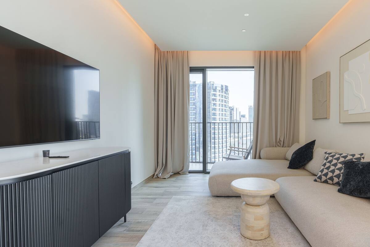 A modern hotel room with a large TV, a sofa, a coffee table, and a balcony with city views.