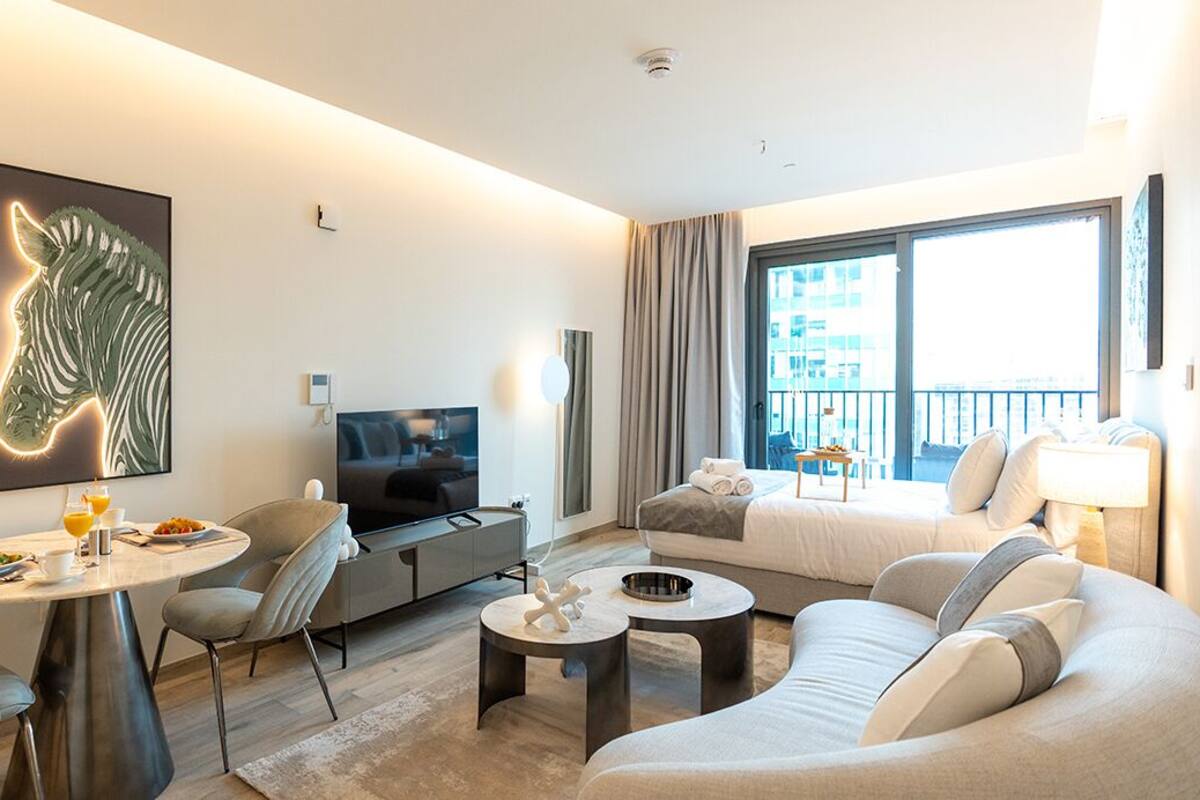 A modern hotel room with a large window, a flat-screen TV, a dining area with a table and chairs, a sofa, and a zebra-themed artwork on the wall.