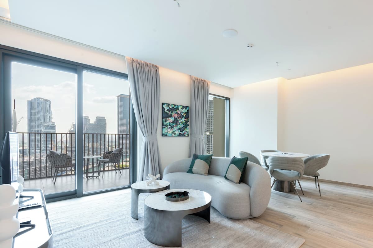 A modern living room with a large sofa, a round coffee table, and a dining area with a small table and chairs. A balcony with a view of the city skyline is visible through large windows.