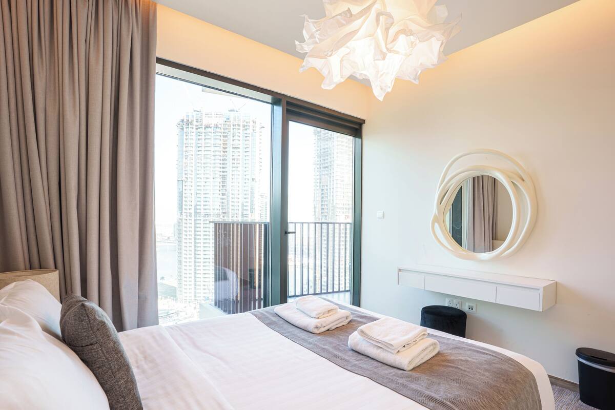 A hotel room with a large bed, a view of tall buildings, a round mirror, and a modern light fixture.