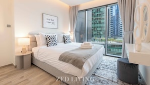 A modern hotel room with a large bed, a bedside table with a lamp, a large window with a view of buildings, and a framed quote on the wall. - Ahad Residences (Dubai)