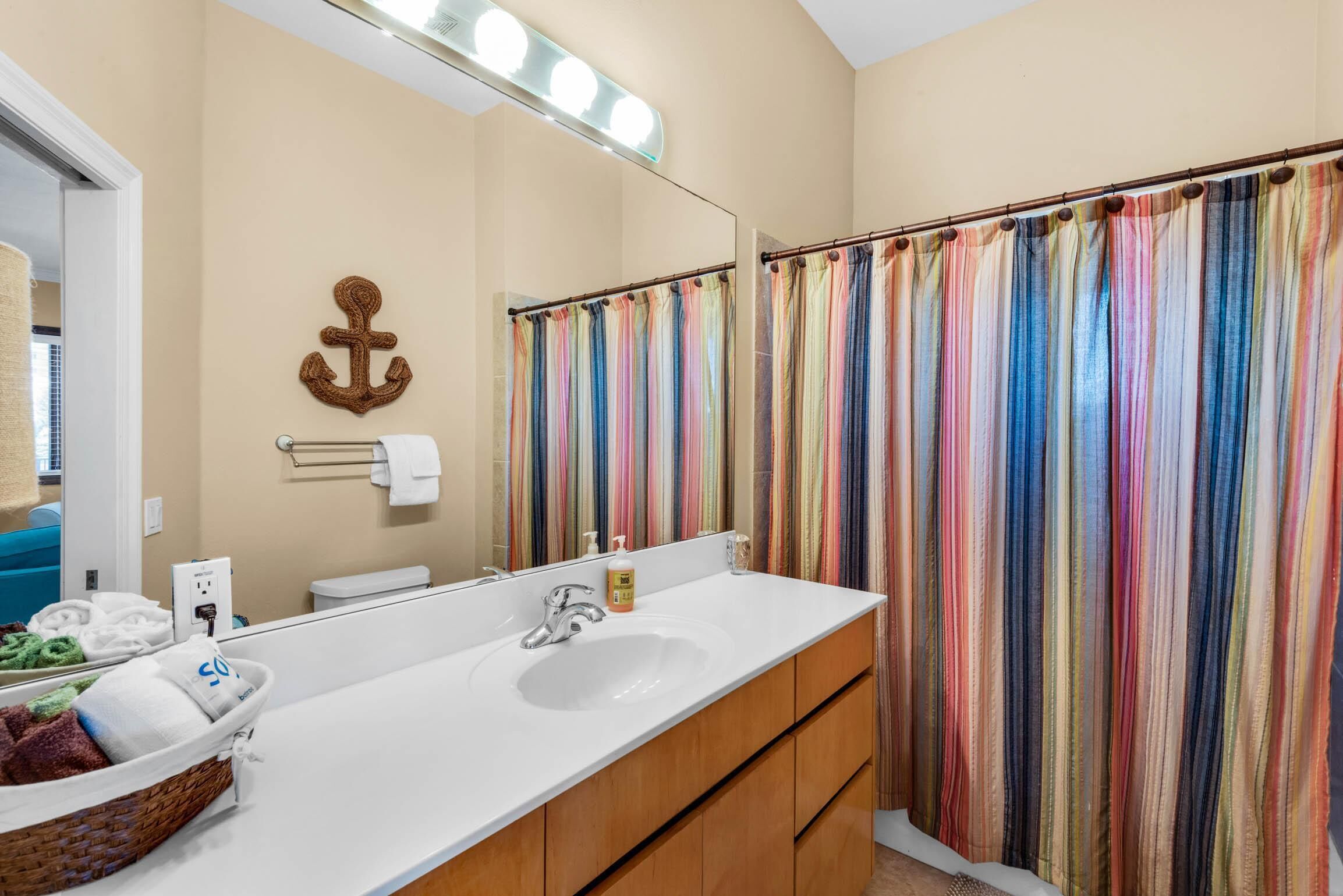 A bathroom with a white sink, wooden cabinets, a mirror with lights, a towel rack, and a colorful striped shower curtain.