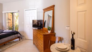 A bedroom with a bed, a dresser, a TV, a mirror, a dining table with plates and a wine bottle, and a sliding door leading to a balcony. - Studio at the PGA (Port St. Lucie)