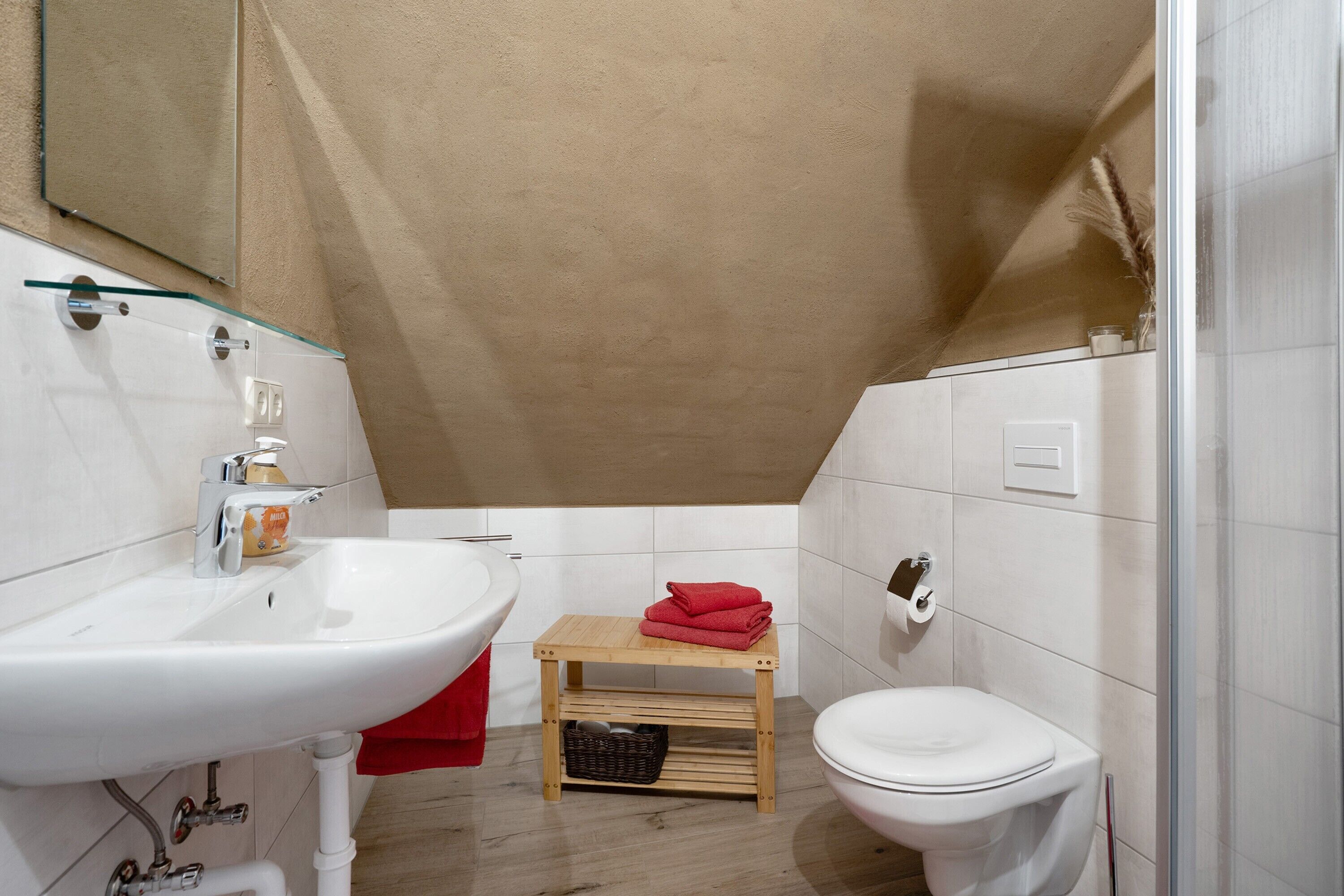 Un baño con un lavamanos blanco, un inodoro, una pequeña mesa de madera con una toalla roja y un estante de vidrio sobre el lavamanos.