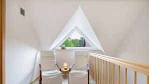 A room with a sloped ceiling, two white chairs, a small table with a lamp, and a window with a view of trees. - Holiday Home 'Bauernnest' with Shared Garden and Wi-Fi (Schmogrow-Fehrow)