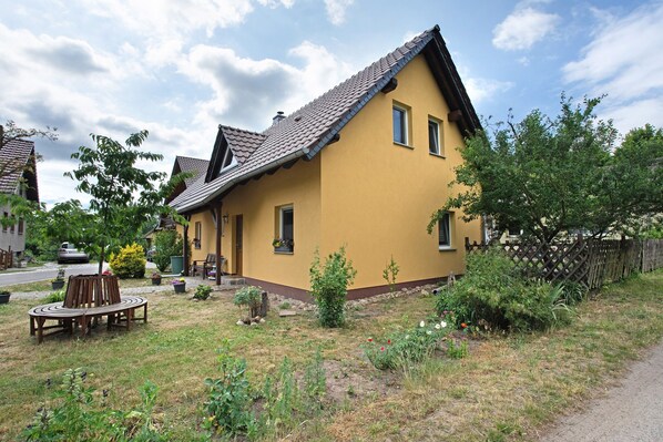 A yellow house with a sloped roof, a garden with various plants, a wooden bench, and a fence. - Holiday Home 'Bauernnest' with Shared Garden and Wi-Fi (Schmogrow-Fehrow)