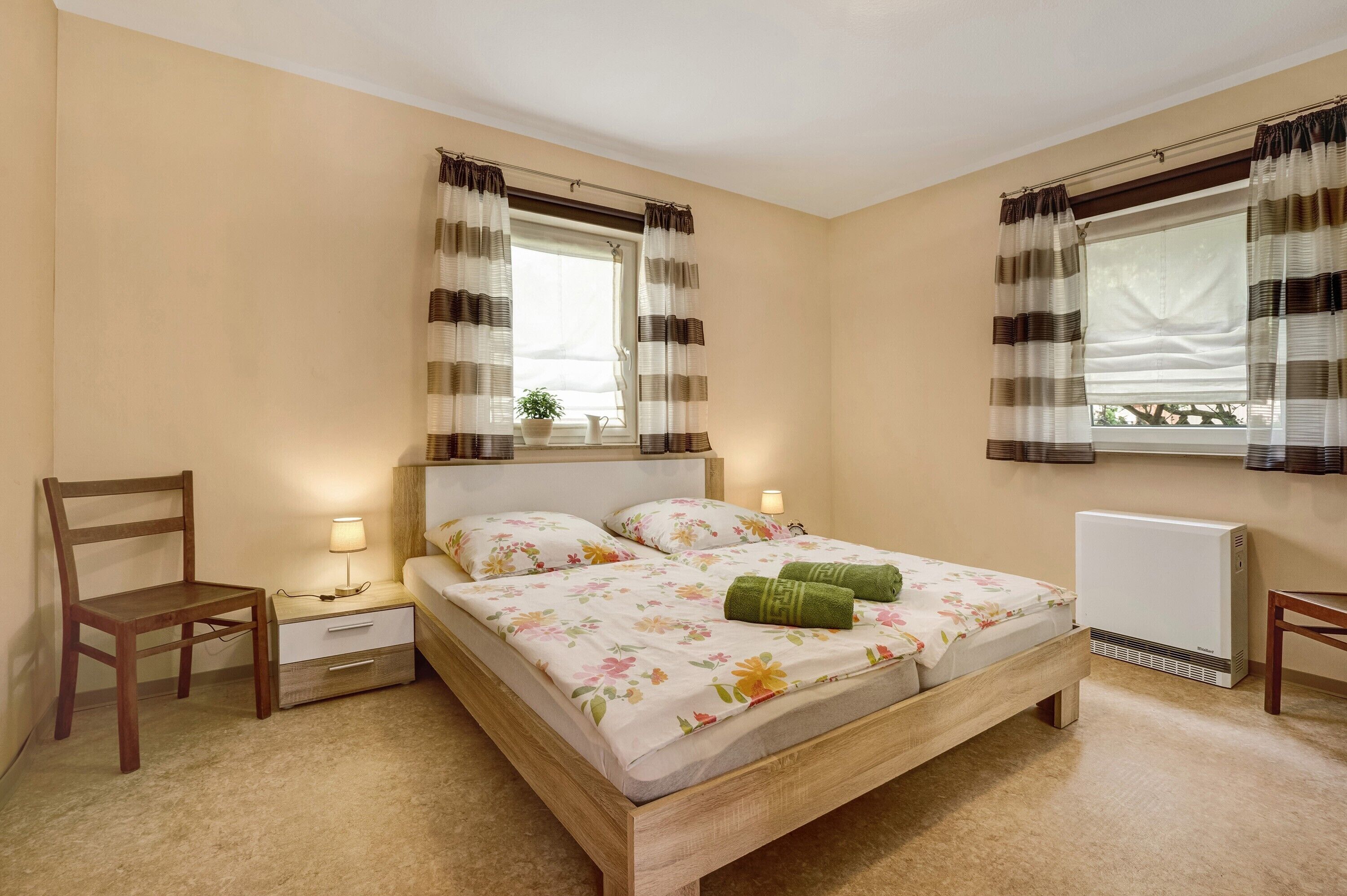Een slaapkamer met een houten bed, twee nachtkastjes met lampen, een stoel, een radiator en een raam met gordijnen.