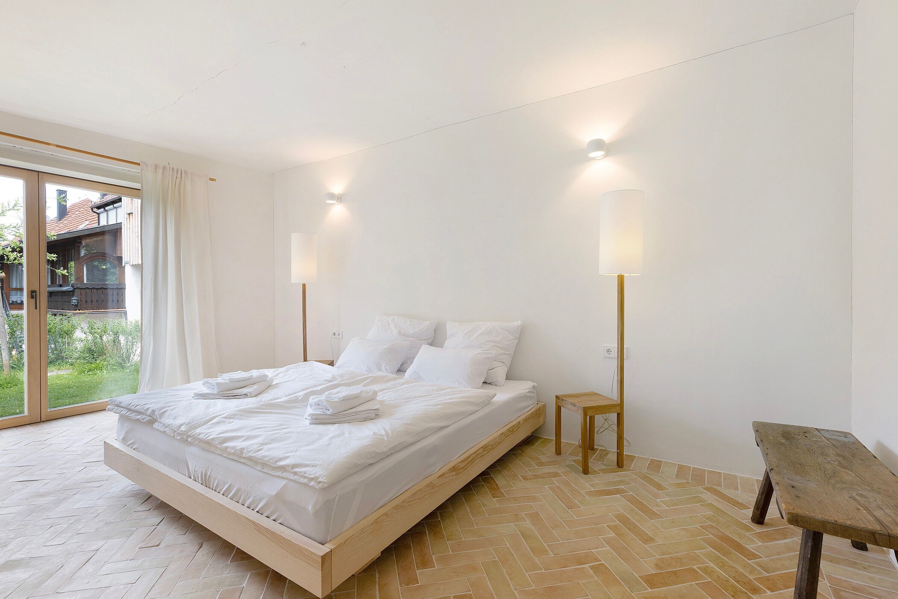 A bedroom with a bed, a wooden bench, a small table, and a view of greenery through a glass door.