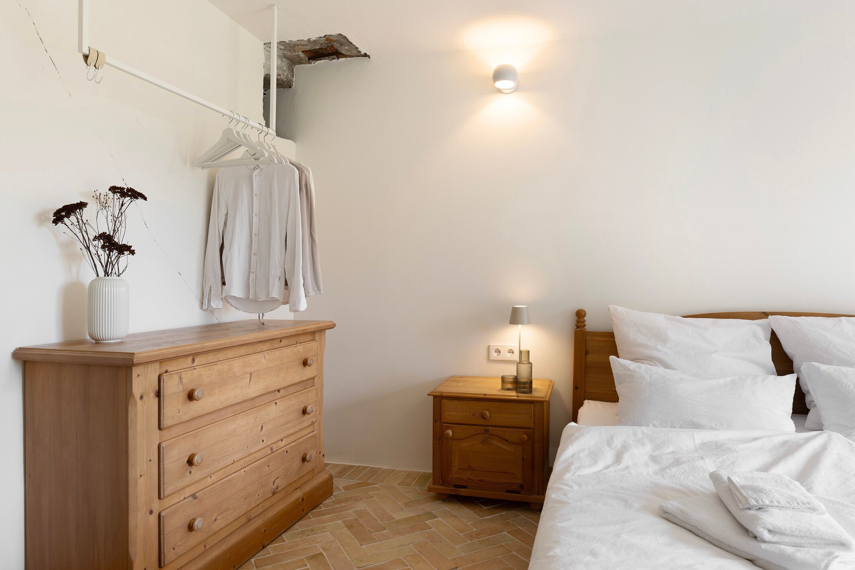 Ein Schlafzimmer mit einem hölzernen Schminktisch, einem Nachttisch mit Lampe, einem Bett mit weißer Bettwäsche und einer Wandleuchte.