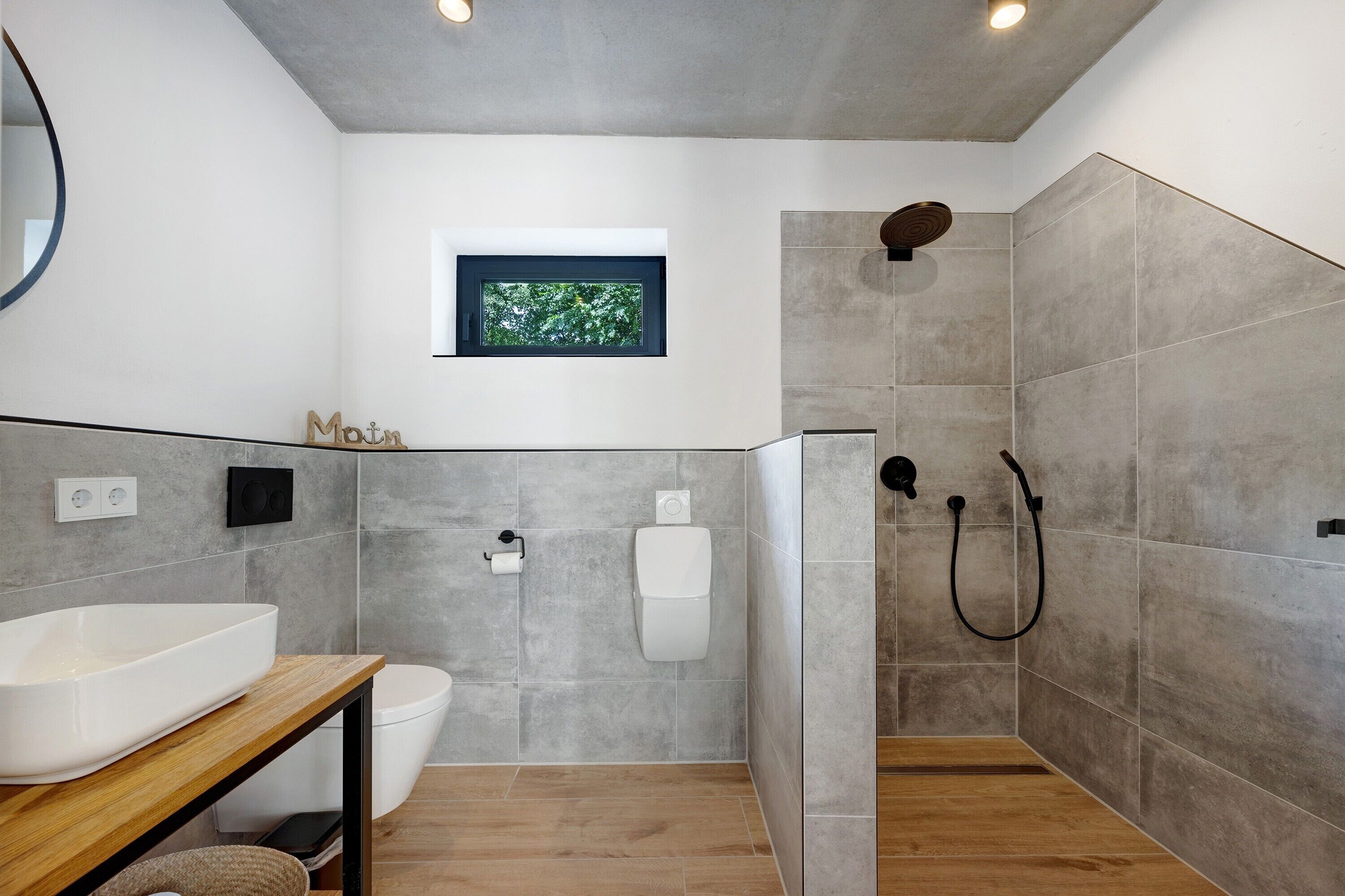 Ein modernes Badezimmer mit einem großen weißen Waschbecken, einer Toilette, einer Duschkabine und einem kleinen Fenster mit Blick auf Bäume.