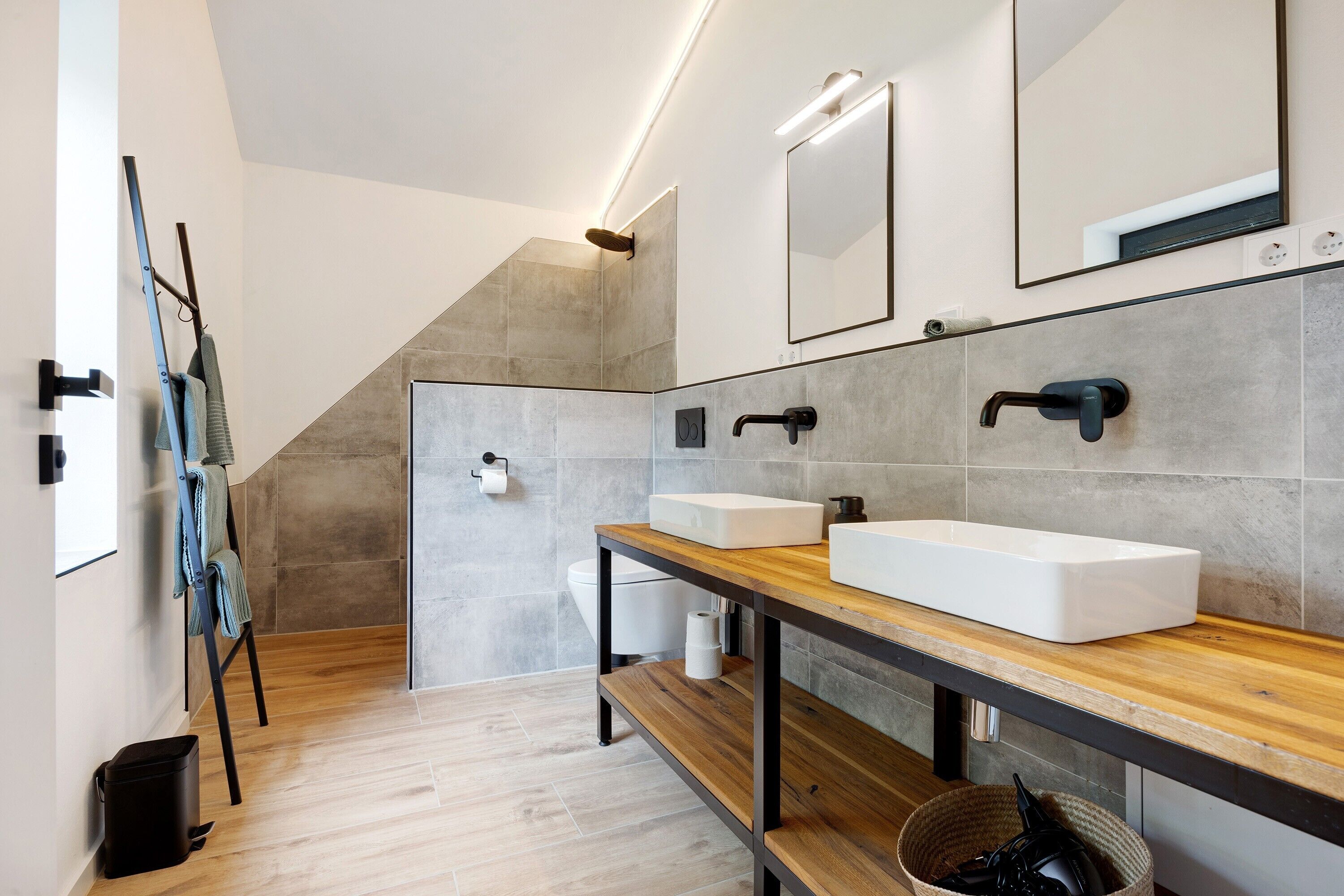 A modern bathroom with two sinks, a wooden vanity, a towel rack, and a shower area.