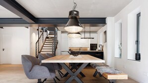 A modern dining area with a wooden table, grey upholstered chairs, a hanging pendant light, and a staircase in the background.