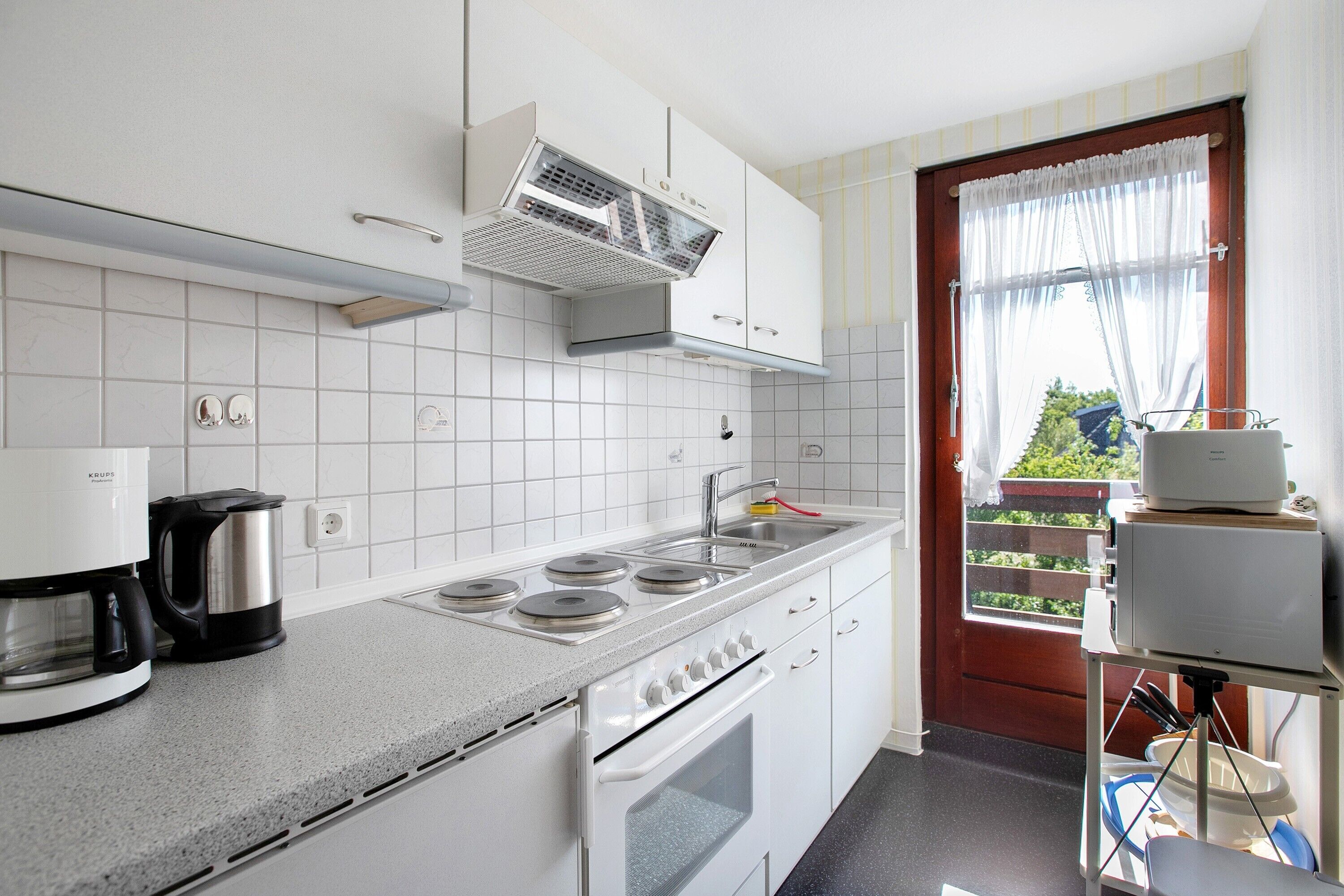 Une cuisine avec des meubles blancs, une cuisinière, une machine à café et une fenêtre avec des rideaux.