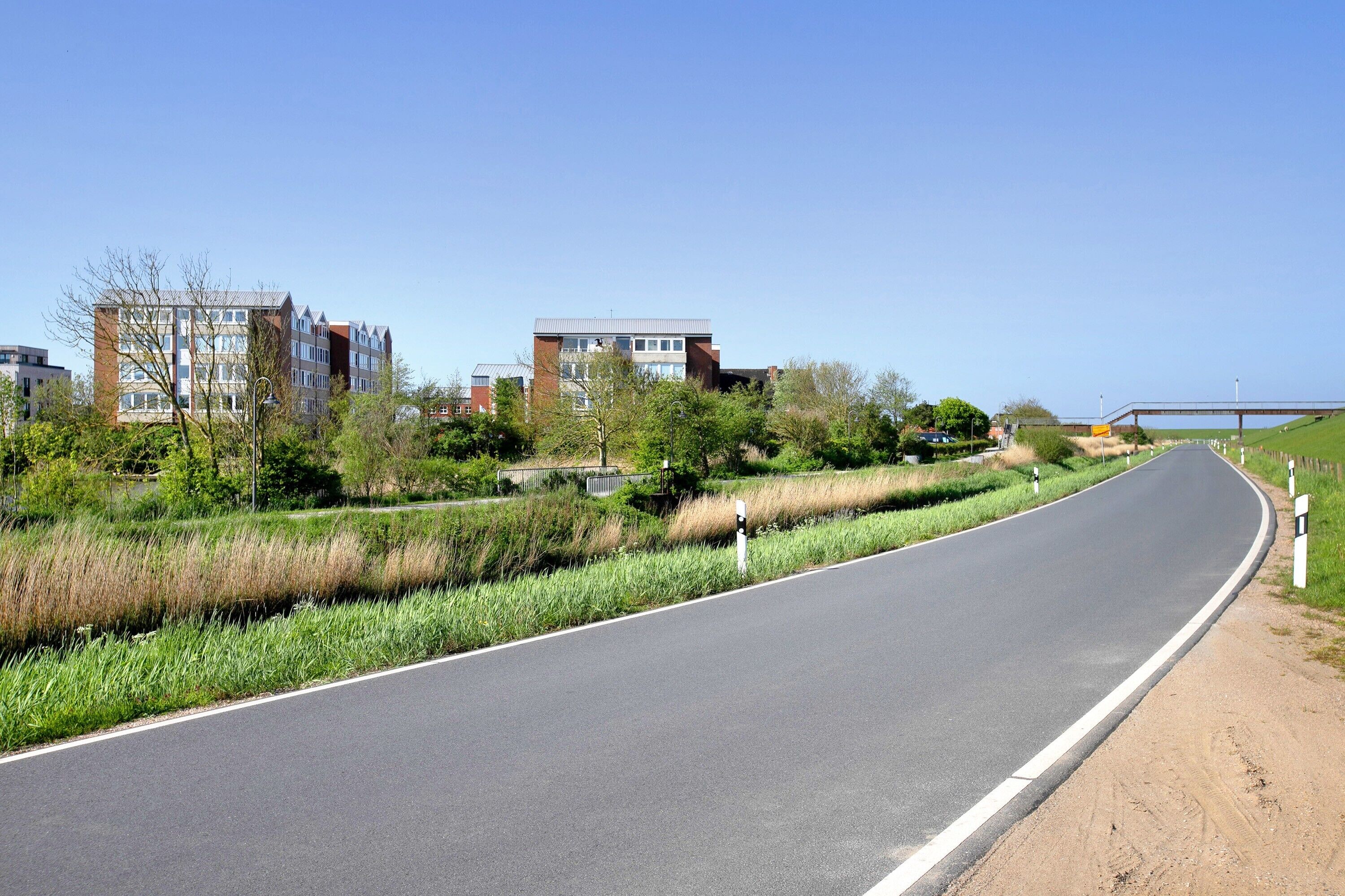 A curved road with buildings in the background, a clear sky, and a grassy area alongside the road.