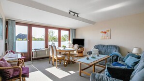 En stue med sofa, spisebord, stole, fjernsyn og et stort vindue.