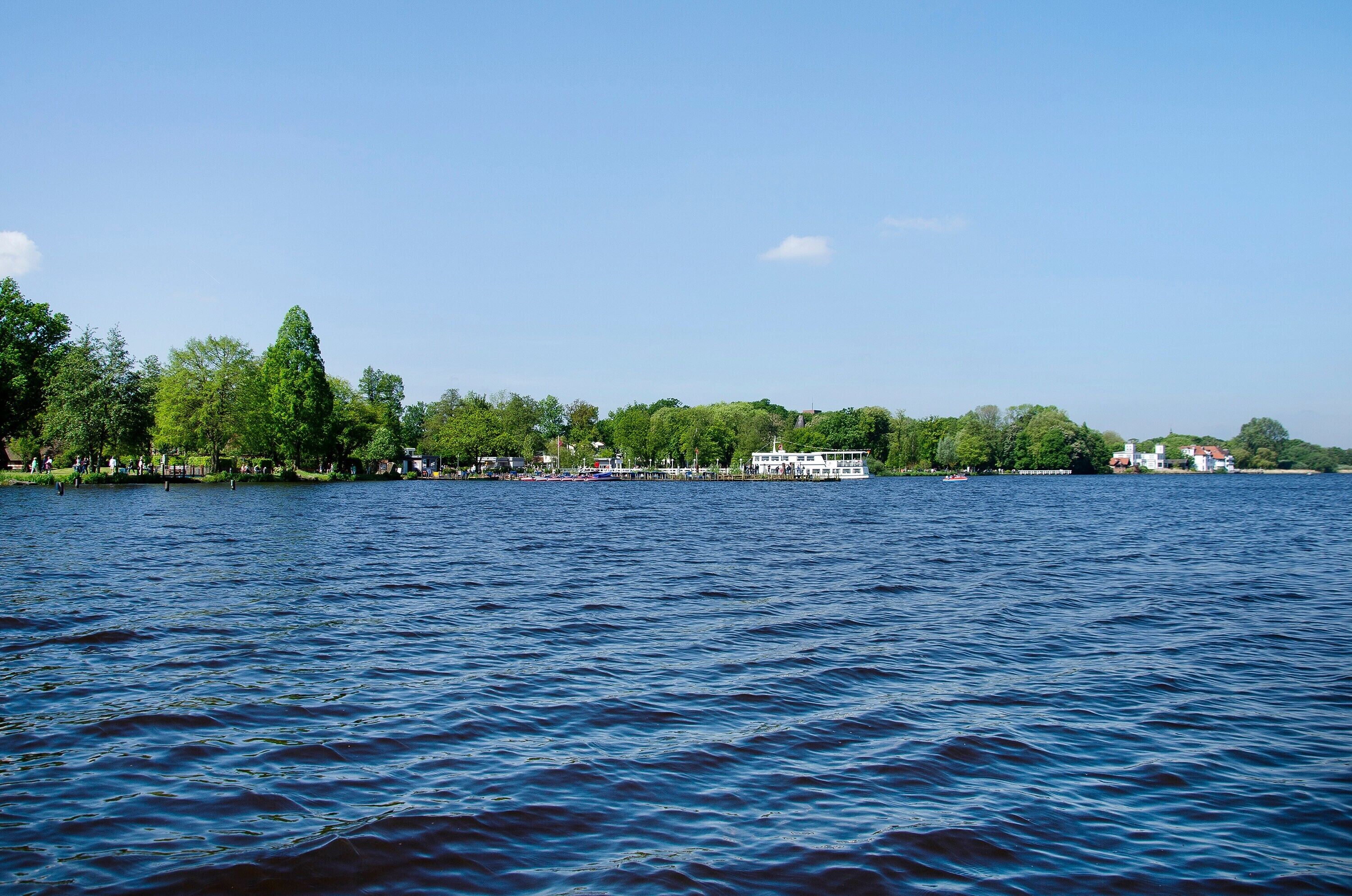 Un lago calmo con una barca ormeggiata a destra, circondato da alberi ed edifici.