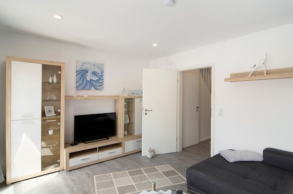 A modern living room with a flat-screen TV, wooden entertainment unit, grey sofa, and a door leading to another room.