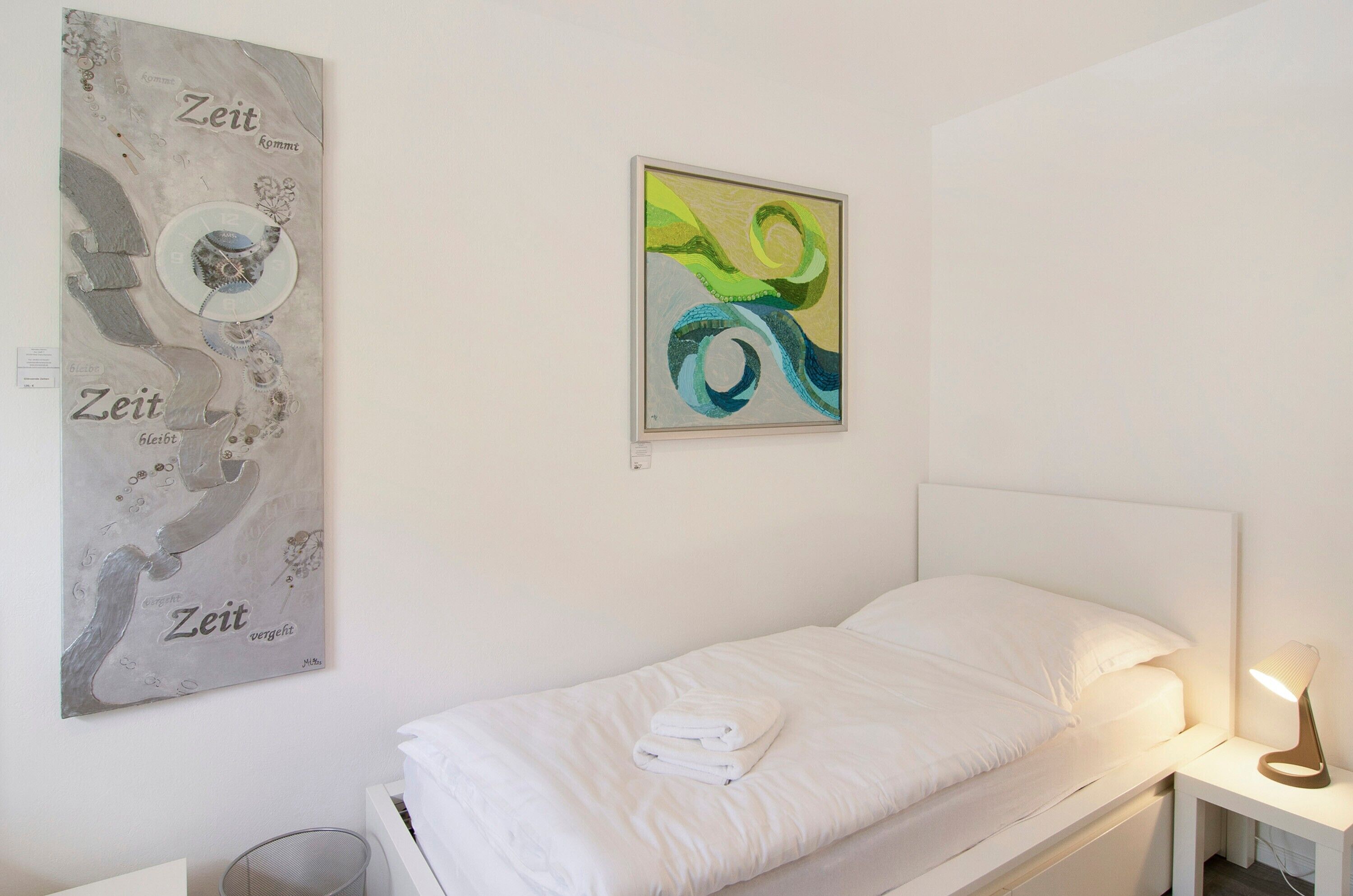 Un lit simple avec une literie blanche, une table de chevet avec une lampe et deux œuvres d’art accrochées au mur.