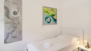 Un lit simple avec une literie blanche, une table de chevet avec une lampe et deux œuvres d’art accrochées au mur.