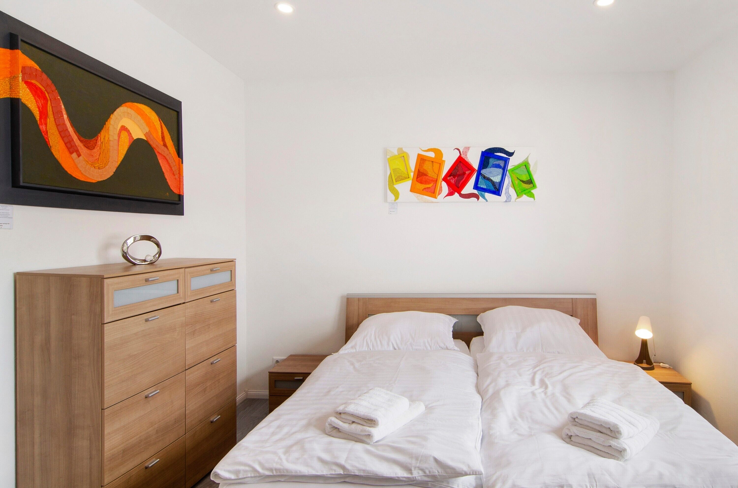 A hotel room with a bed, a wooden dresser, a nightstand with a lamp, and two wall art pieces.