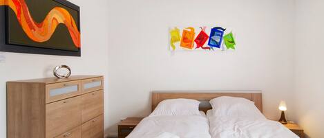 A hotel room with a bed, a wooden dresser, a nightstand with a lamp, and two wall art pieces.