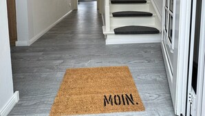Un pasillo con una puerta de madera, una escalera y un felpudo que dice 'MOIN'.