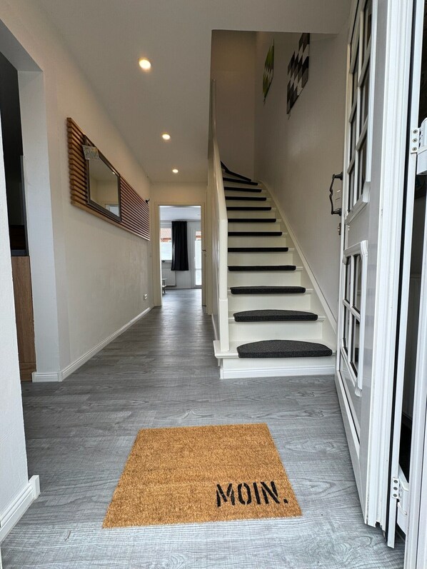 A hallway with a wooden door, a staircase, and a doormat with 'MOIN' on it.