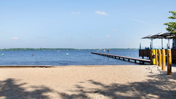 Una playa con un muelle de madera que se adentra en aguas tranquilas, una orilla arenosa y una pequeña construcción con techo.