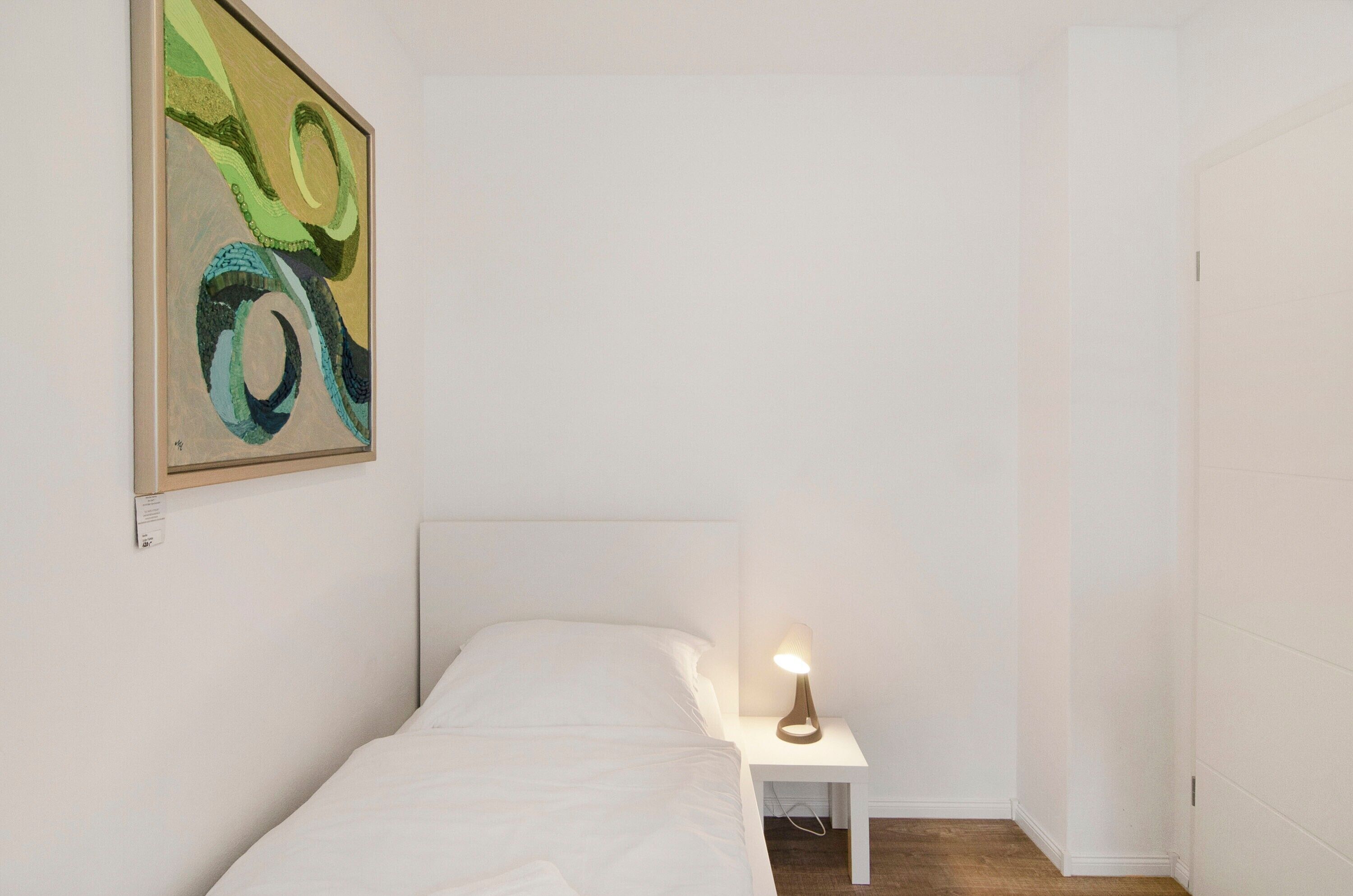 A single bed with a white headboard, a bedside table with a lamp, and a wall-mounted abstract painting.