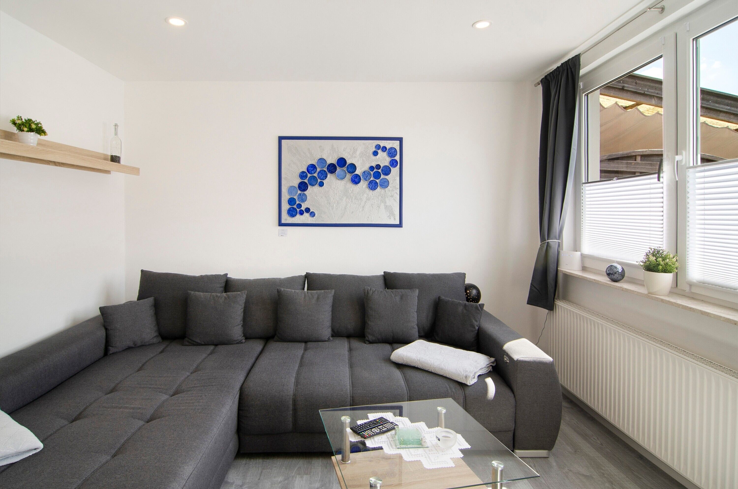 Ein modernes Wohnzimmer mit grauem Sofa, einem Glastisch und einem blauen, abstrakten Gemälde an der Wand.