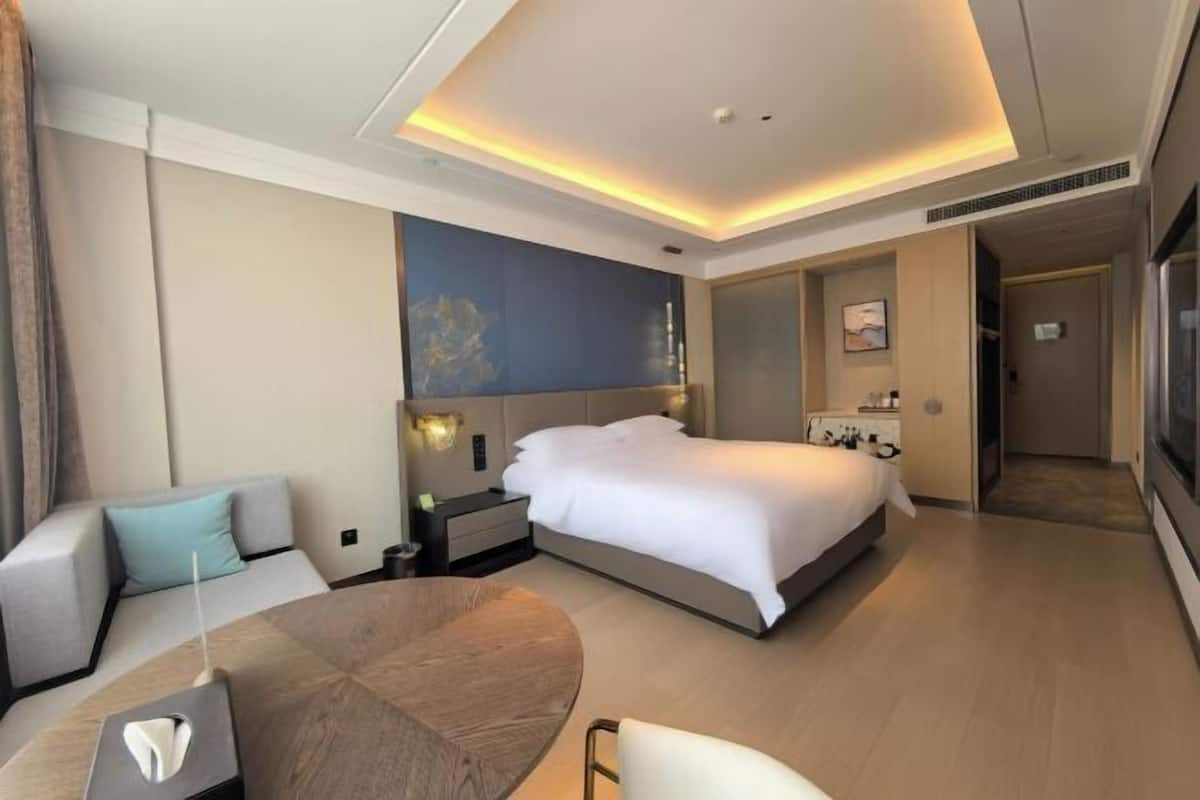Ein modernes Hotelzimmer mit einem großen Bett, einem runden Esstisch, einer Couch und einem durch eine offene Tür einsehbaren Badezimmer.