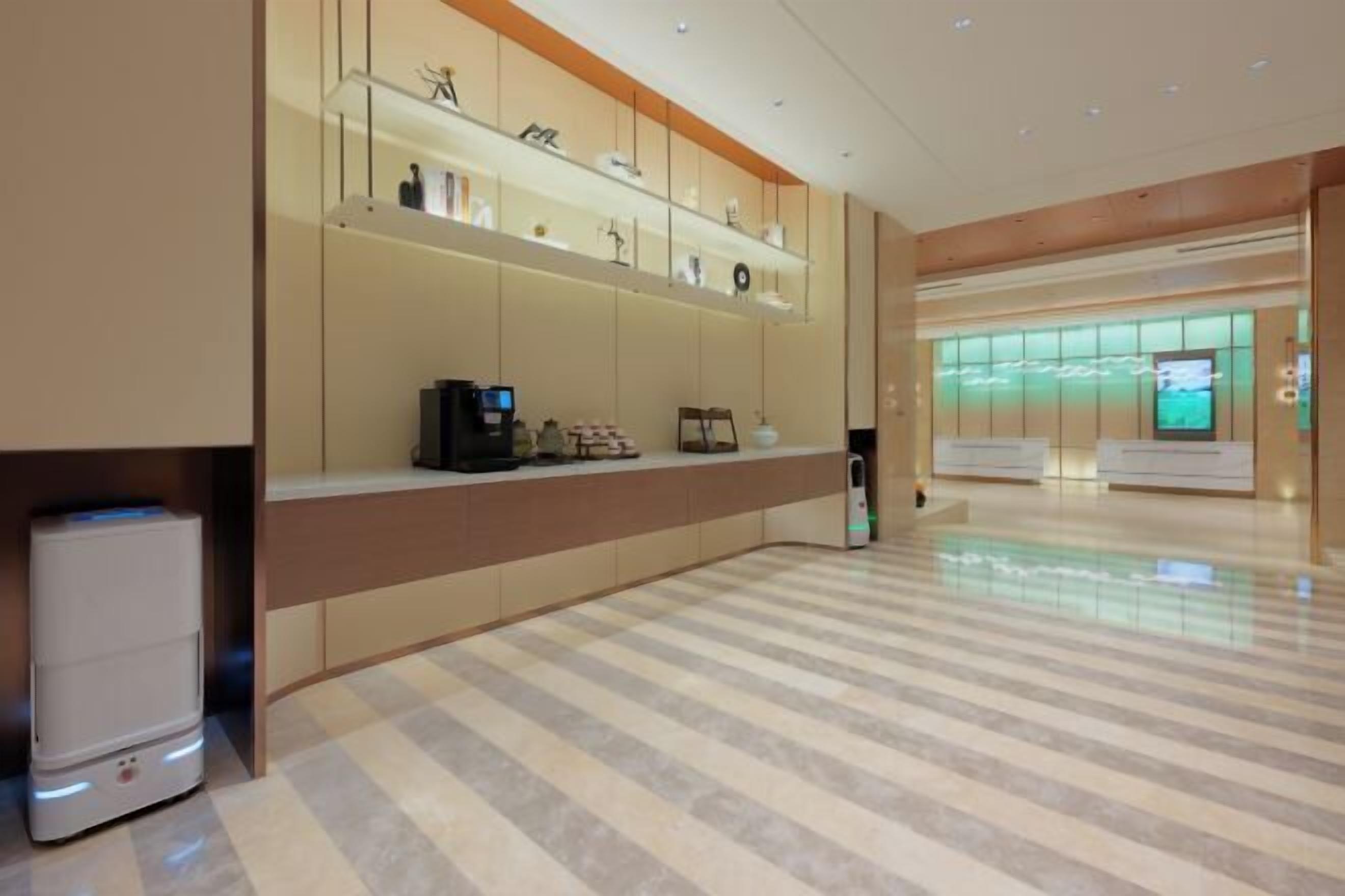 Un moderno vestíbulo de hotel con mostrador de recepción, una máquina de café y un área de estar con una pared iluminada en verde.