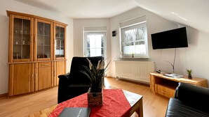 A living room with a wooden cabinet, a black chair, a red table with a book, a plant, and a TV mounted on the wall.