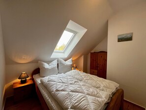 Une chambre avec un plafond en pente, un puits de lumière, un lit avec une literie blanche, une table de chevet en bois avec une lampe et une armoire en bois.