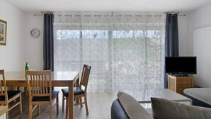 Una habitación con una mesa de comedor, sillas, un sofá, un televisor sobre un mueble de madera y una ventana con cortinas translúcidas.