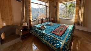 A bedroom with a wooden ceiling, a bed with a patterned bedspread, a chair, and a nightstand with a lamp. - SUNS Cottage (Kasauli)