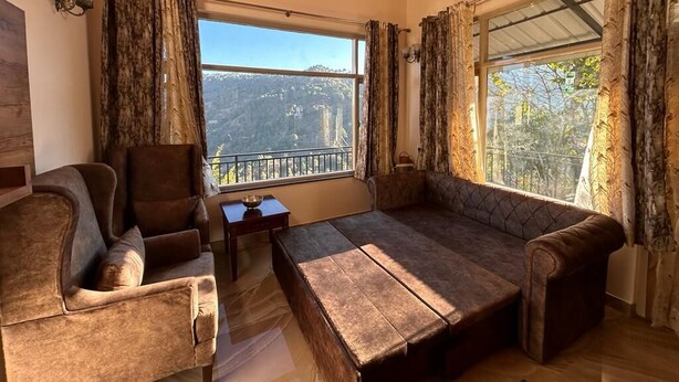 A cozy room with a sofa and armchair, large windows with curtains, and a view of a mountainous landscape. - SUNS Cottage (Kasauli)