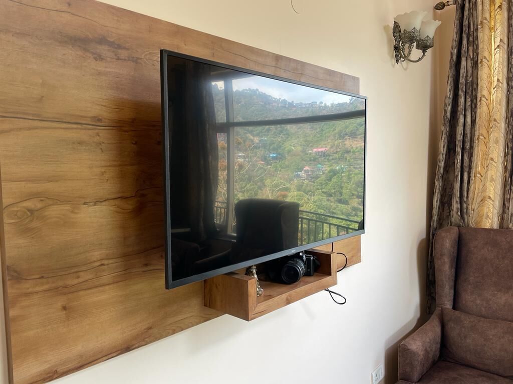 A flat-screen TV mounted on a wooden wall with a camera below it, a brown armchair to the right, and a scenic view of a hilly landscape through the TV.