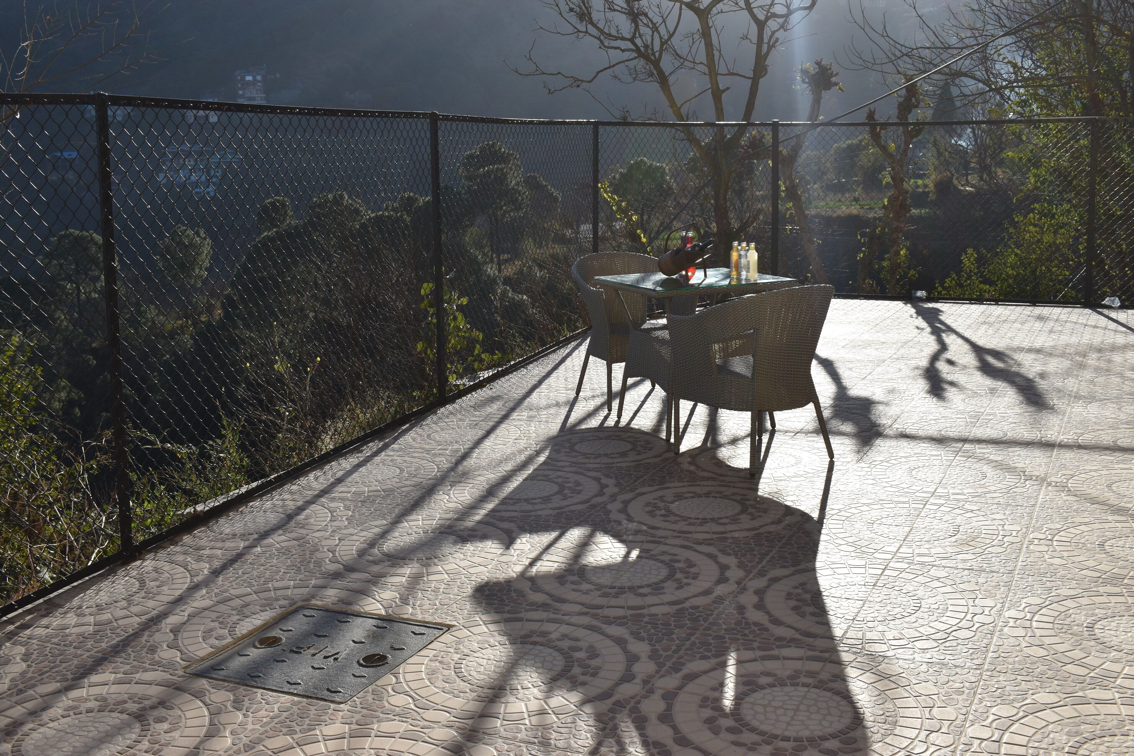 A terrace with a patterned floor, a table with chairs, and a view through a chain-link fence.