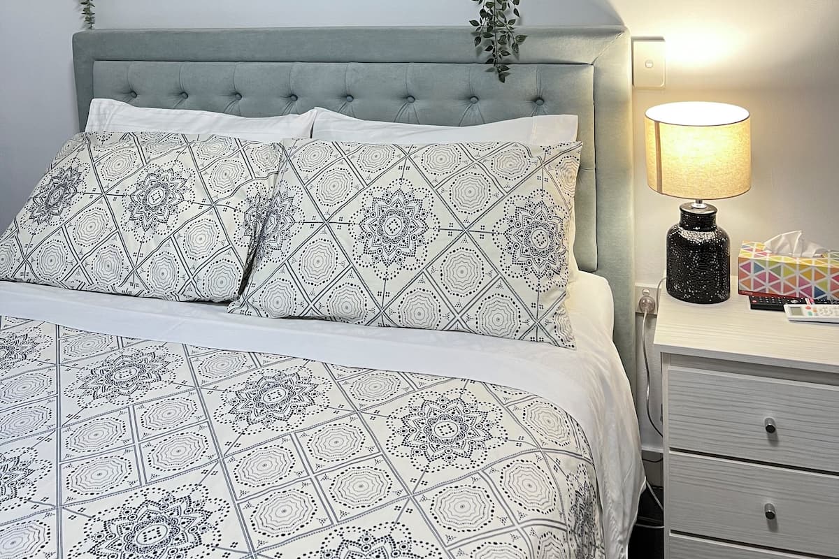 Un dormitorio ordenado con una cama, una mesita de noche con una lámpara y un cuadro en la pared.