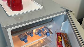Et køleskab med vanddispenser, elkedel og forskellige madvarer, herunder snacks og vand på flaske.