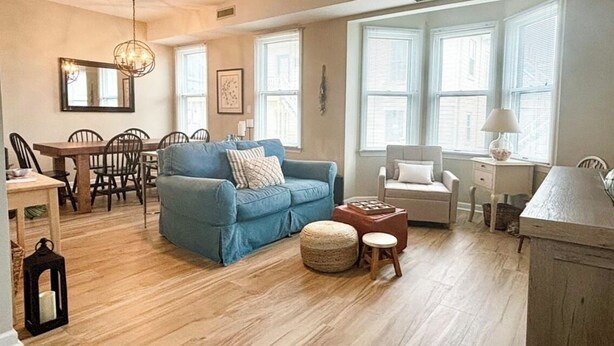 Un salon spacieux avec un canapé bleu, une table à manger avec des chaises, un coin cuisine doté d’un plan de travail blanc et de grandes fenêtres qui laissent entrer la lumière naturelle.