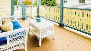 Une véranda couverte avec des meubles en osier et une table, une clôture en bois peinte en jaune et une vue sur les maisons voisines.