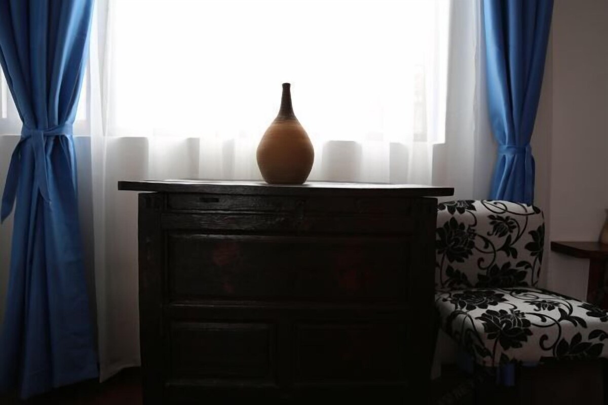 Ein Zimmer mit einem dunklen Holzschrank, einer Vase darauf und einem gemusterten Sessel.