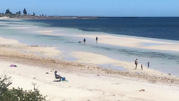 Ein Strand mit klarem Wasser, Menschen, die spazieren gehen, und eine Person, die auf einem Stuhl sitzt.